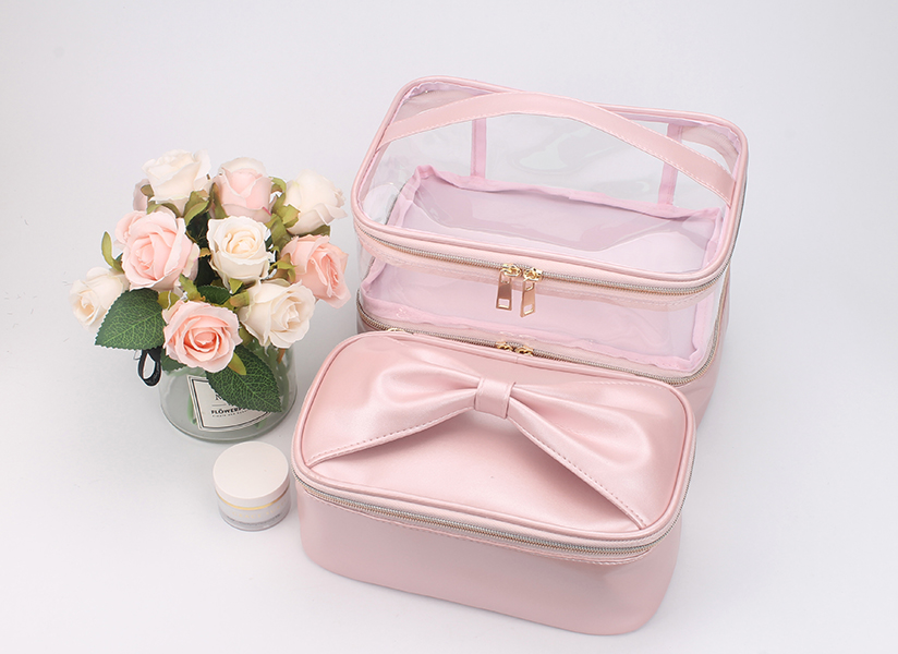 Two-layer pink satin cosmetic bag with transparent PVC upper compartment, bow decoration, top hand strap and metal double zippers